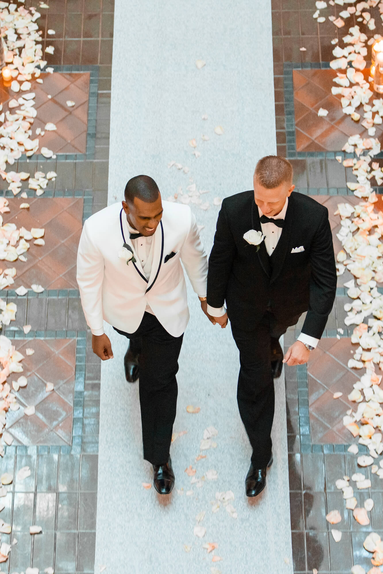 Two men in tuxedos walk down an aisle decorated with flower petals and floral arrangements on sides.