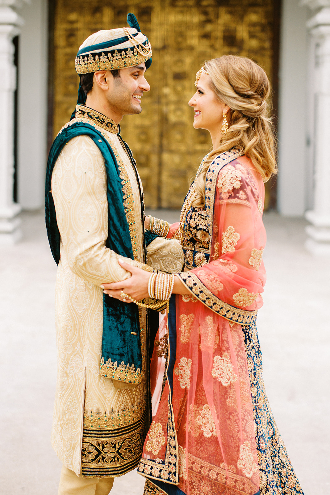 A man and woman in traditional attire face each other, holding hands, smiling, with a classical architectural background.