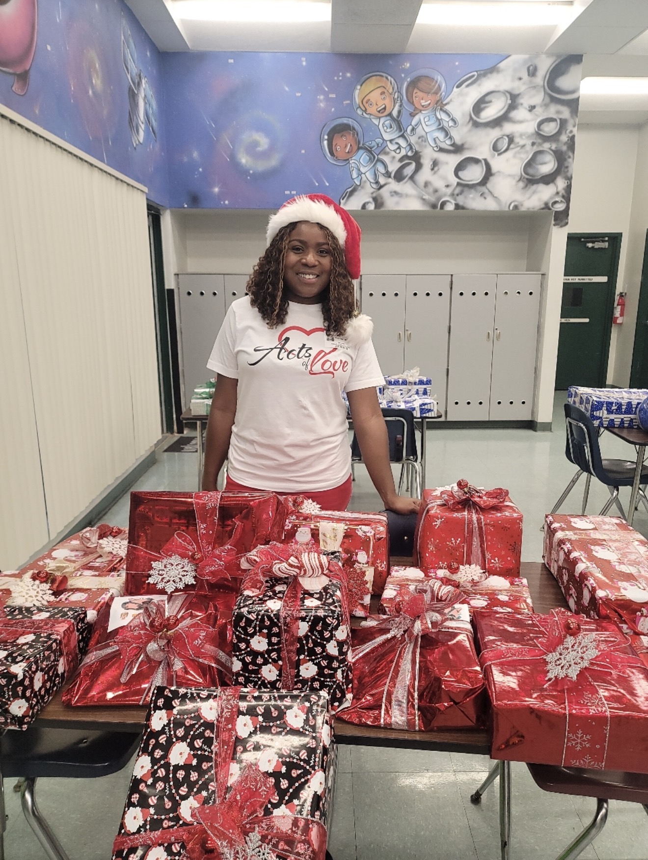 Woman wearing a Santa hat and white t-shirt standing behind a table with wrapped Christmas presents in a decorated room.