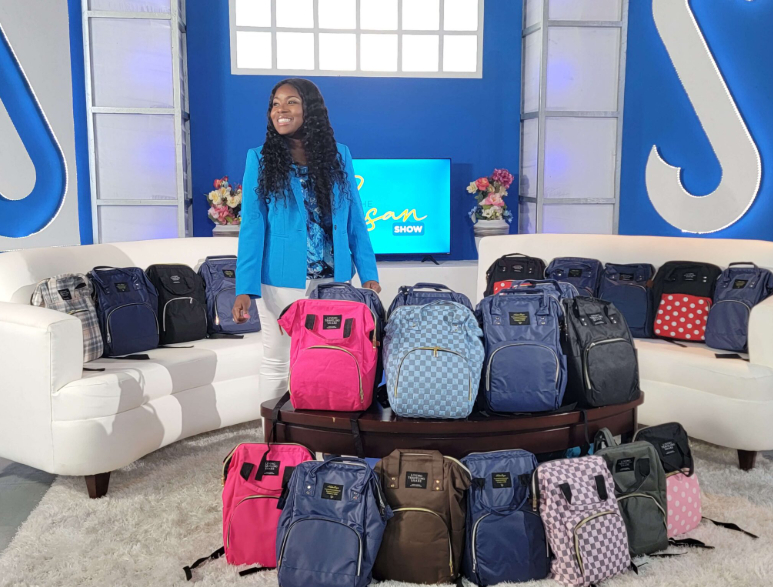Young woman with long curly hair standing behind a table with colorful backpacks, in a bright studio setting.