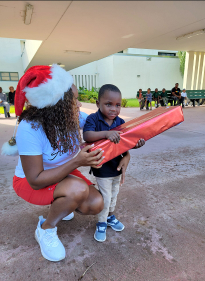 Girl wearing a Santa hat kneeling and handing a wrapped gift to a boy outdoors.