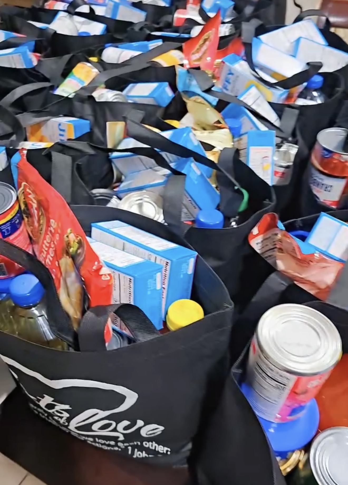 Multiple black bags filled with various food and beverage items, including boxes, bottles, and cans, on a table.