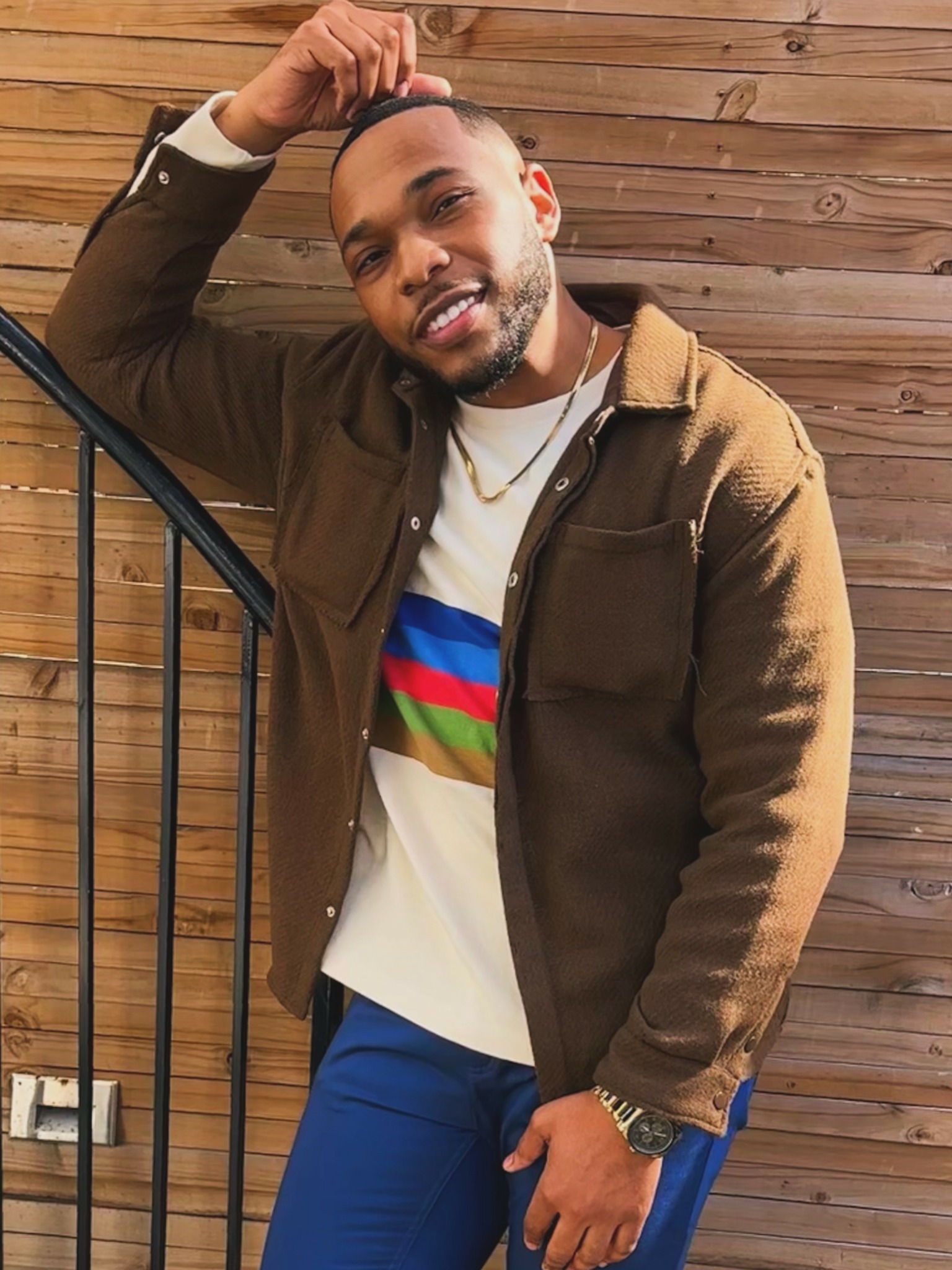 Man smiling, leaning on railing, wooden wall background, casual clothing, gold watch, necklace, short hair, beard.