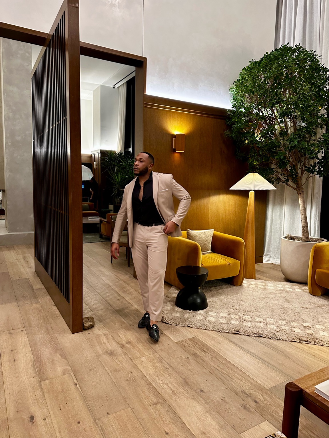 Man in beige suit walking in a modern interior with yellow sofa, large plant, and floor lamp.