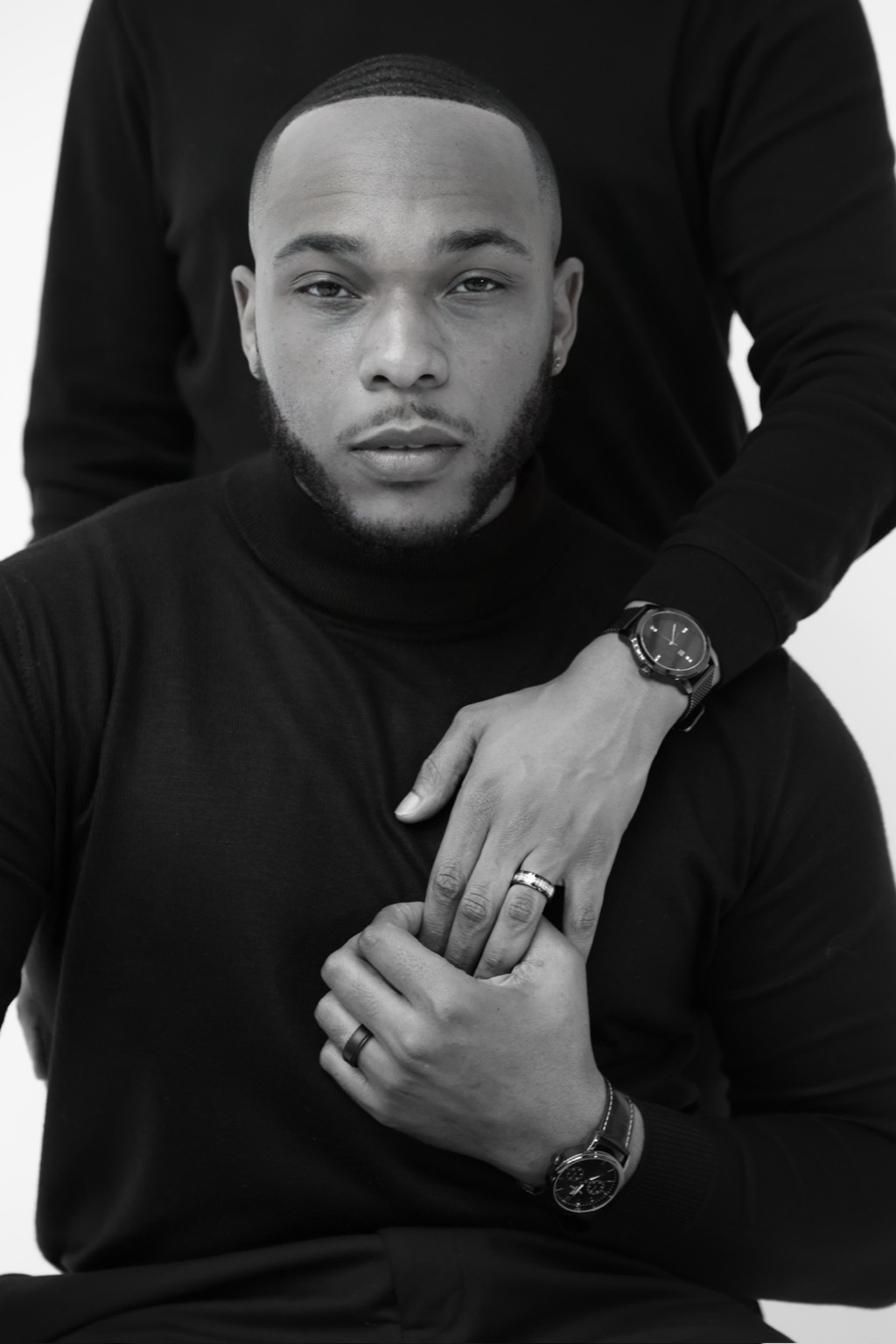 Black and white portrait of a man with short hair, wearing a black shirt, with hands crossed and rings visible.