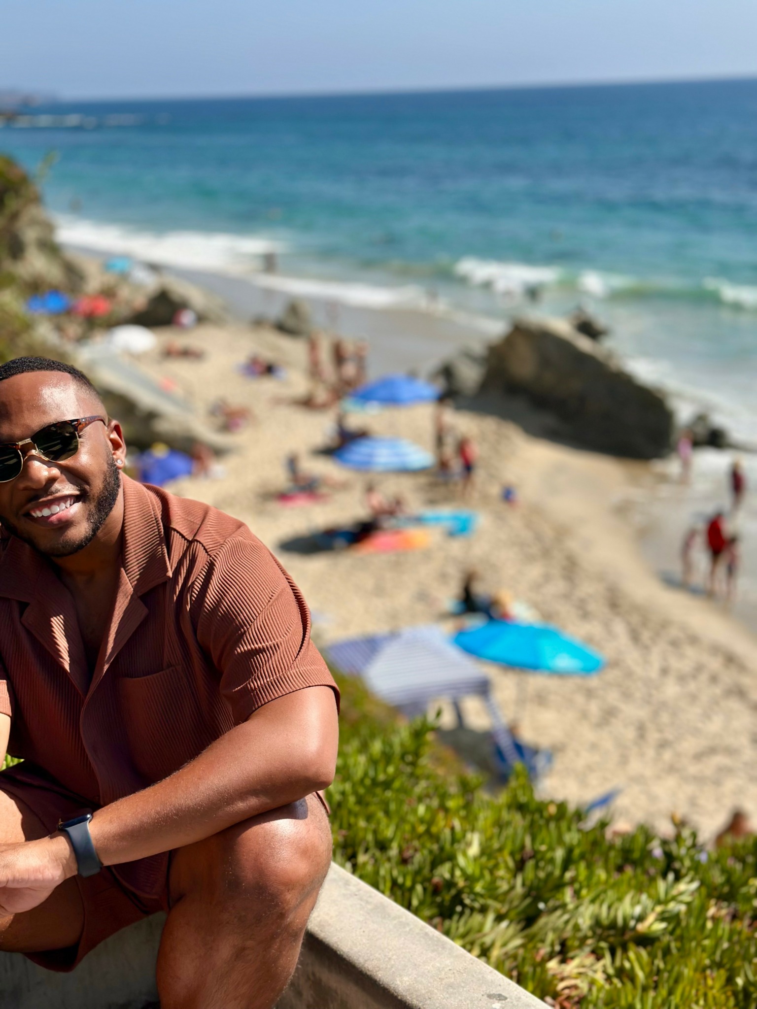 Smiling man wearing sunglasses and a brown shirt sitting outdoors near a beach with umbrellas and rocks.