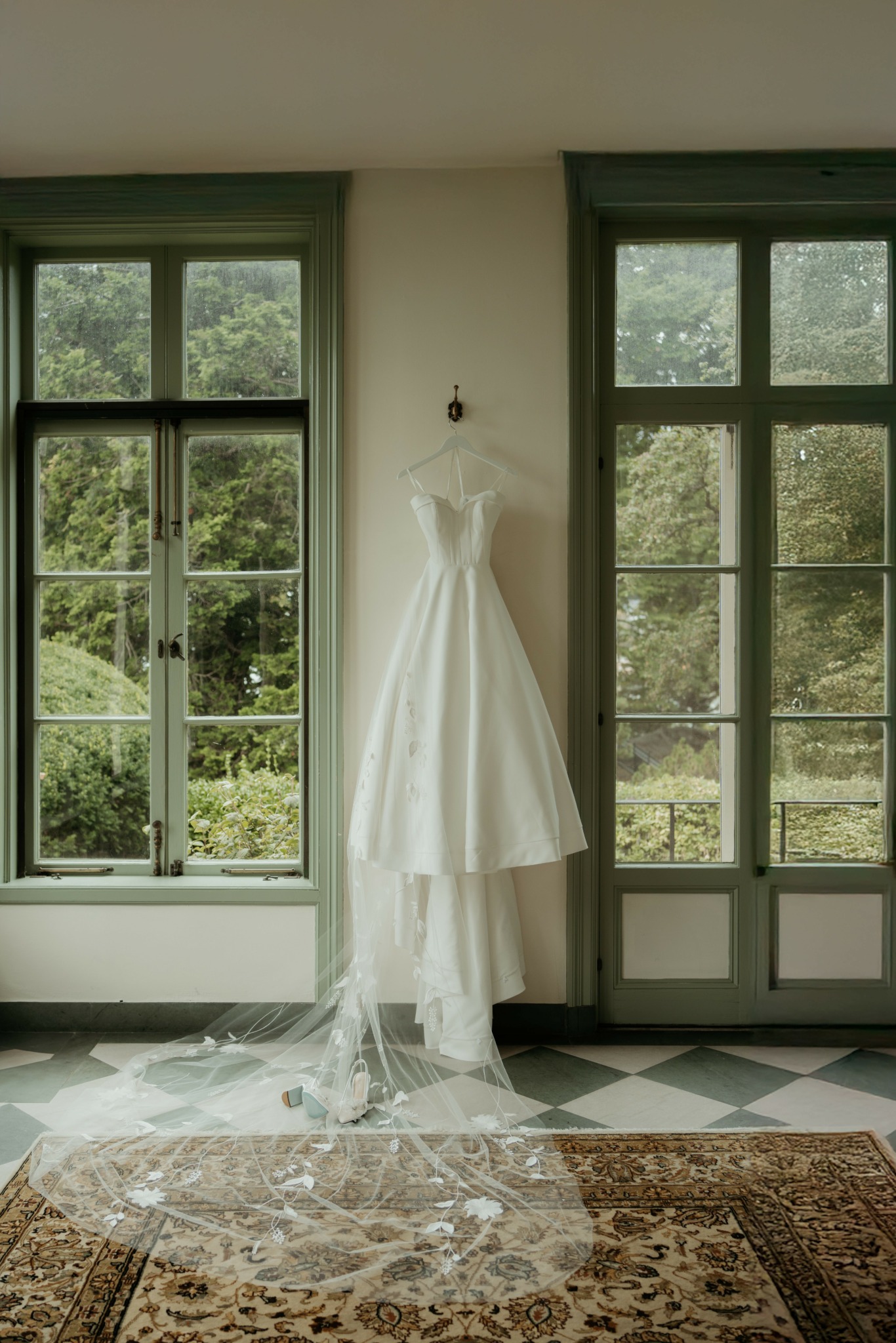 Wedding dress hanging between two large windows with greenery outside, lace veil on the floor, patterned rug below.