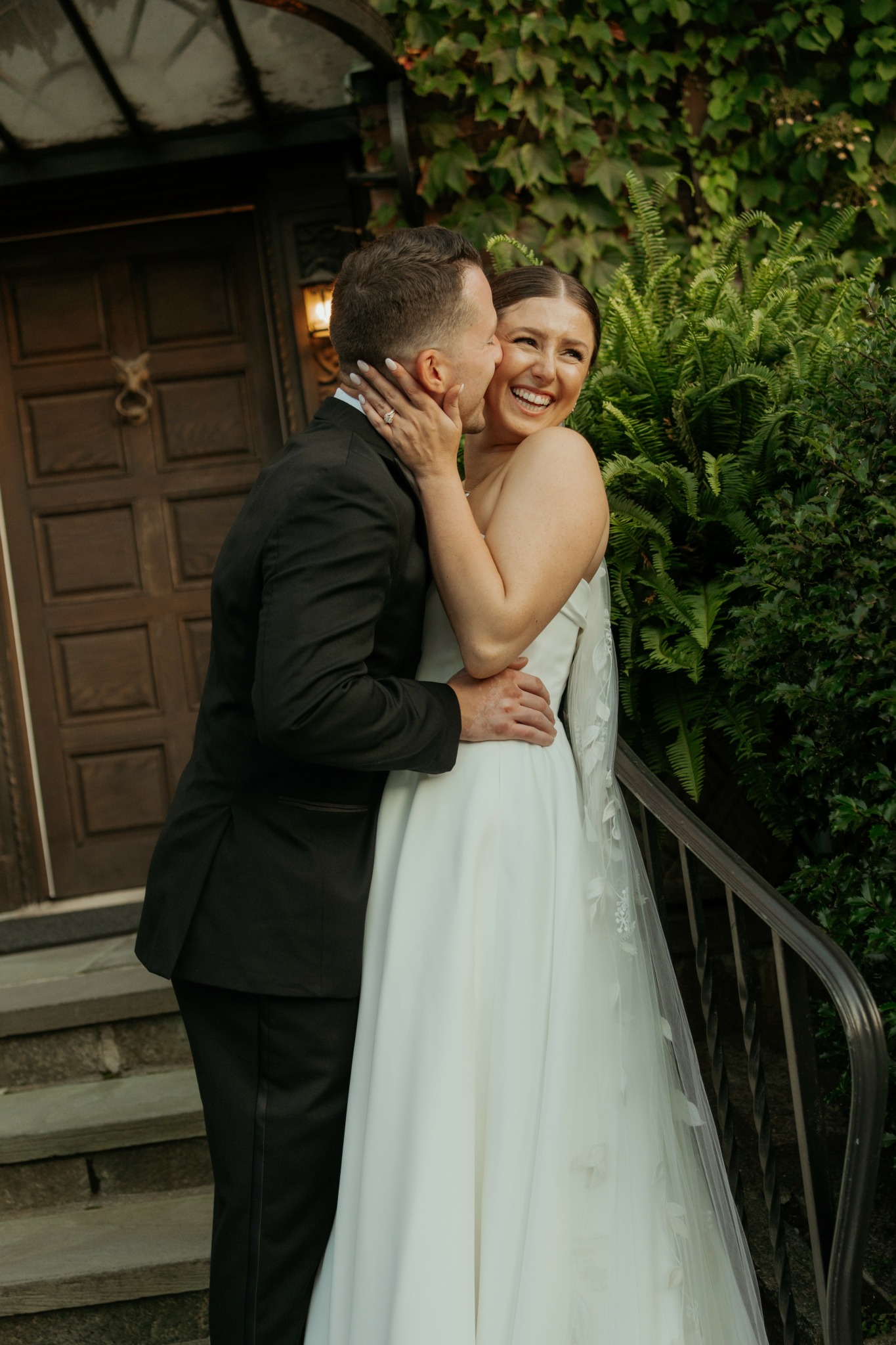A couple in wedding attire sharing a joyful moment outdoors, with greenery and a wooden door in the background.