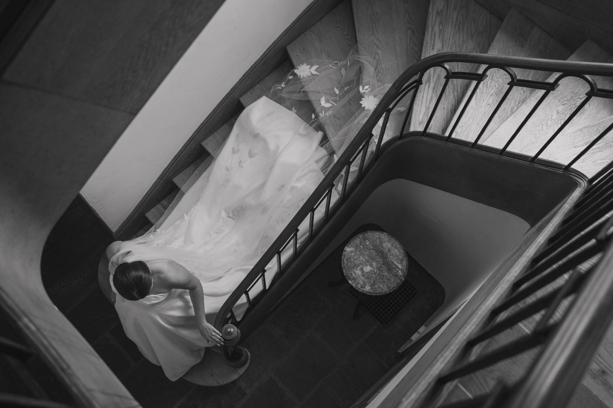 Woman in white dress walking down a staircase with a black railing, viewed from above.
