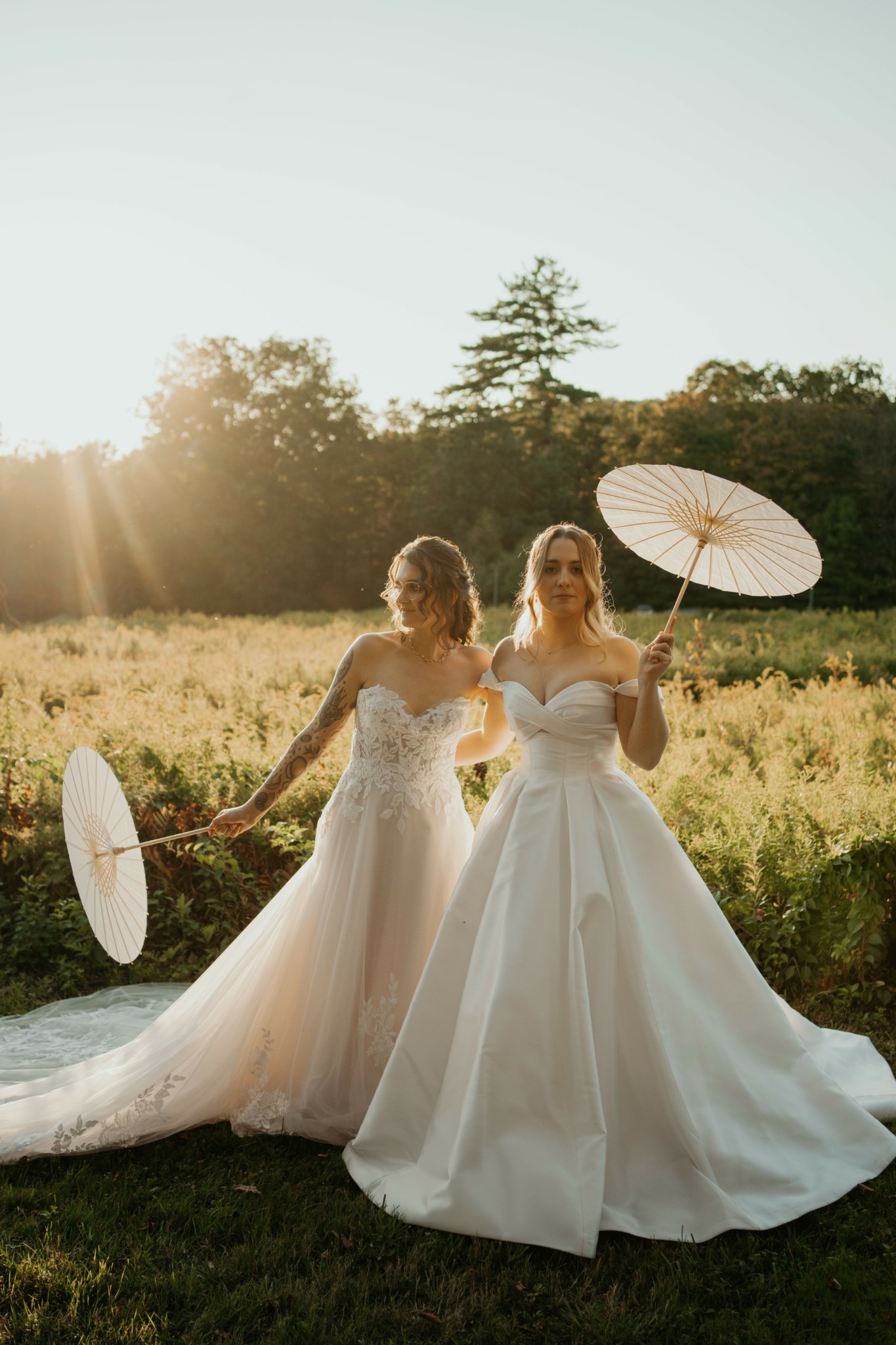 Two women in wedding dresses holding umbrellas in a field with trees and sunlight in the background.