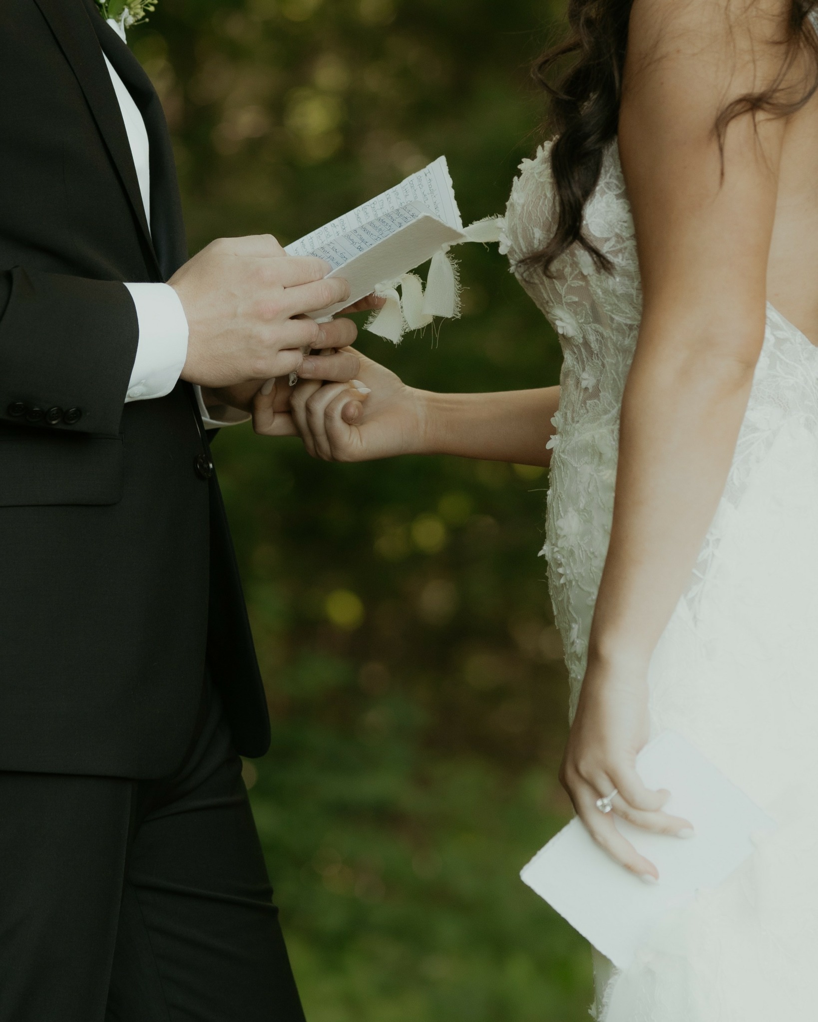 Two people holding hands, one reading from a paper, outdoors with blurred green background.