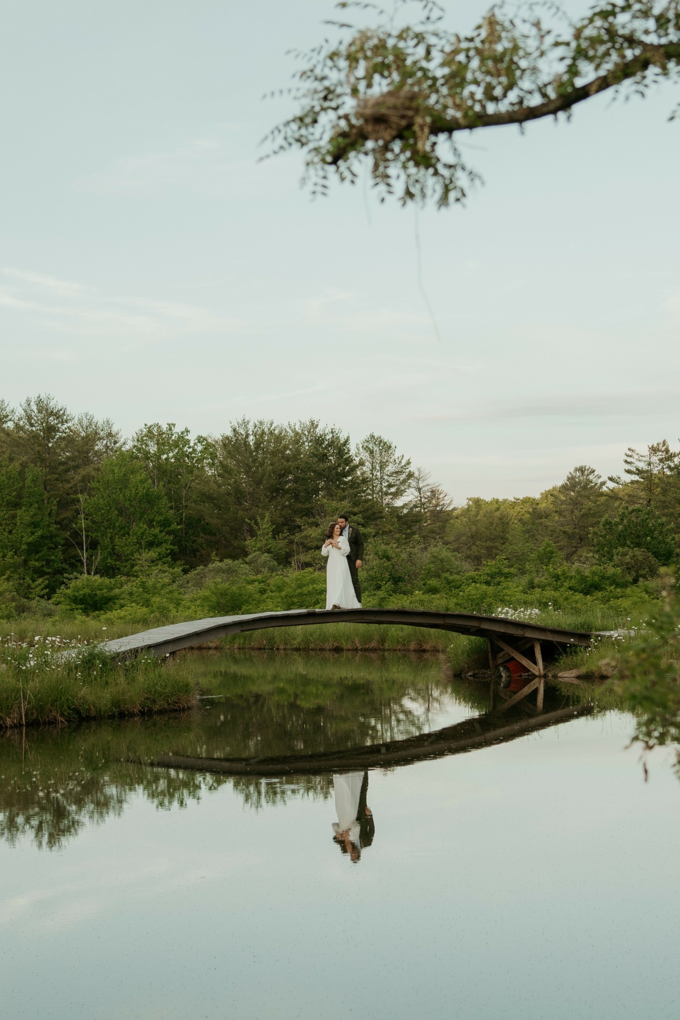 Person standing on a small bridge over a calm body of water, surrounded by trees and greenery.