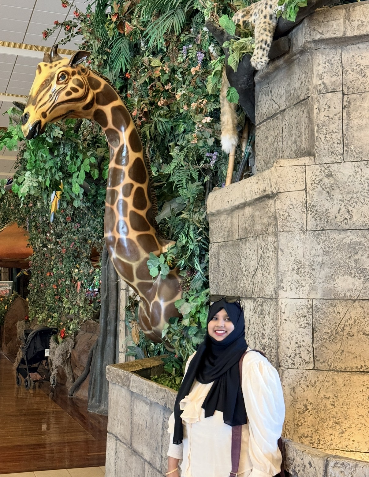 Woman in white and black hijab smiling near a giraffe statue inside a zoo or exhibit.