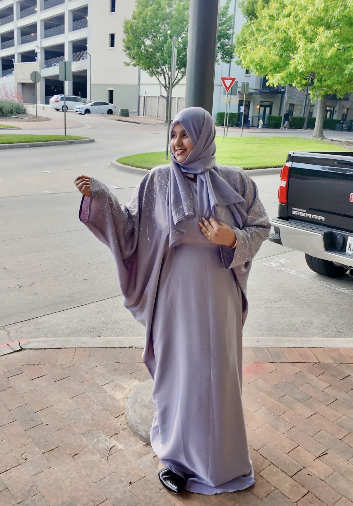 Woman in a long, light purple dress and hijab standing outdoors on brick pavement, smiling, with trees and buildings in background.