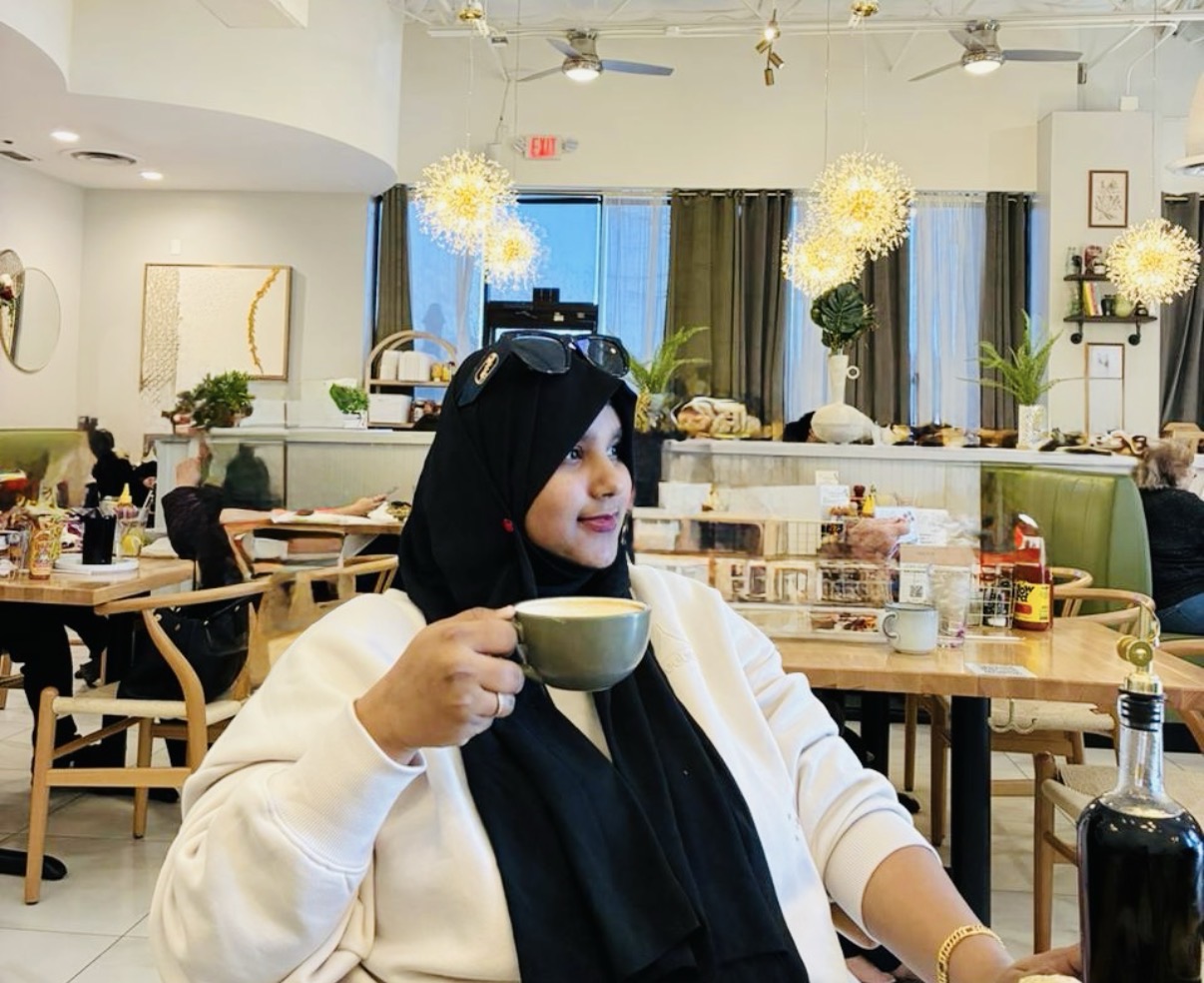 Woman wearing a hijab sitting in a cafe, holding a bowl, with tables, plants, and hanging lights in background.