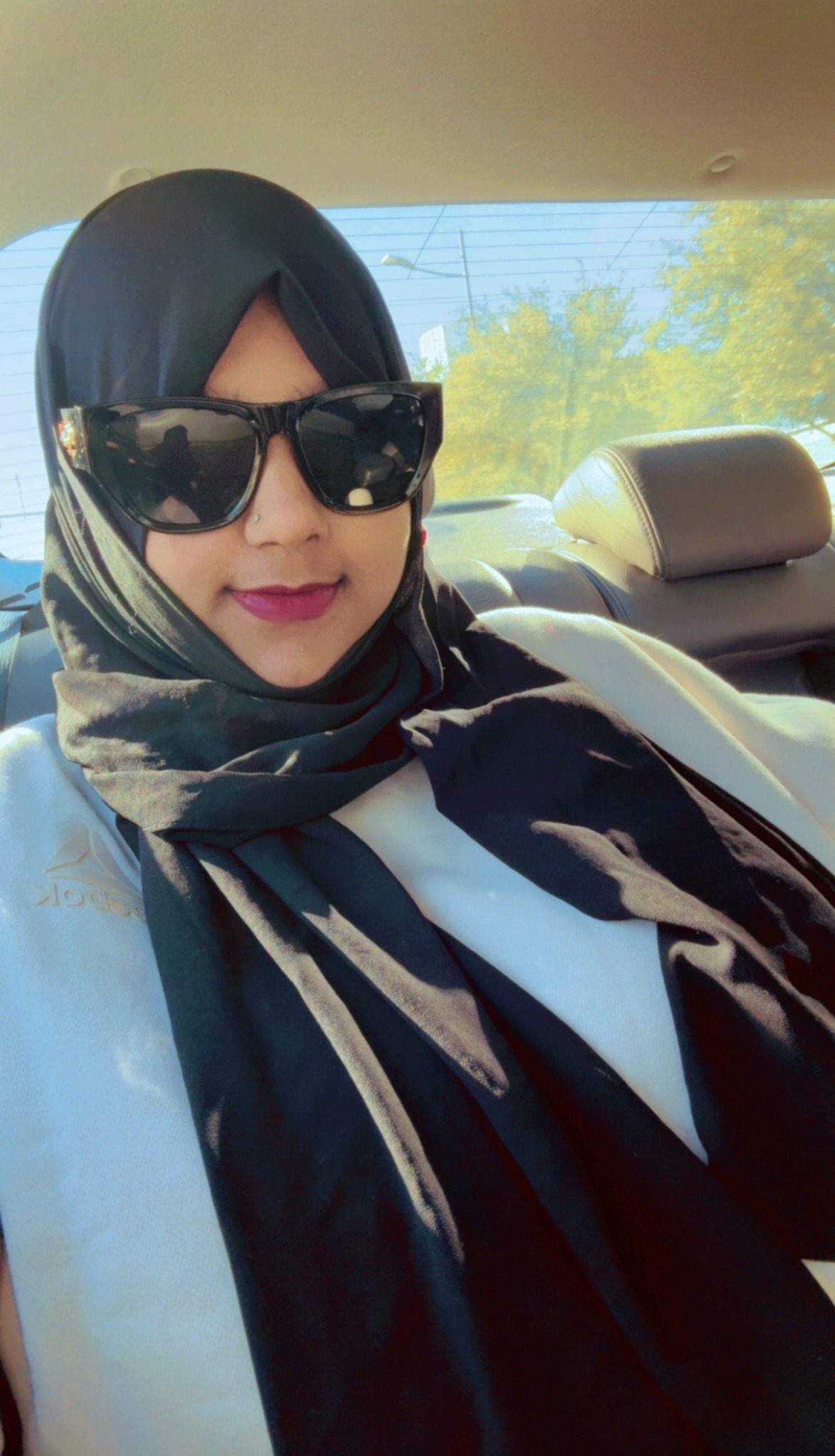 Woman wearing sunglasses and a headscarf, sitting in car, smiling, with trees visible outside window.