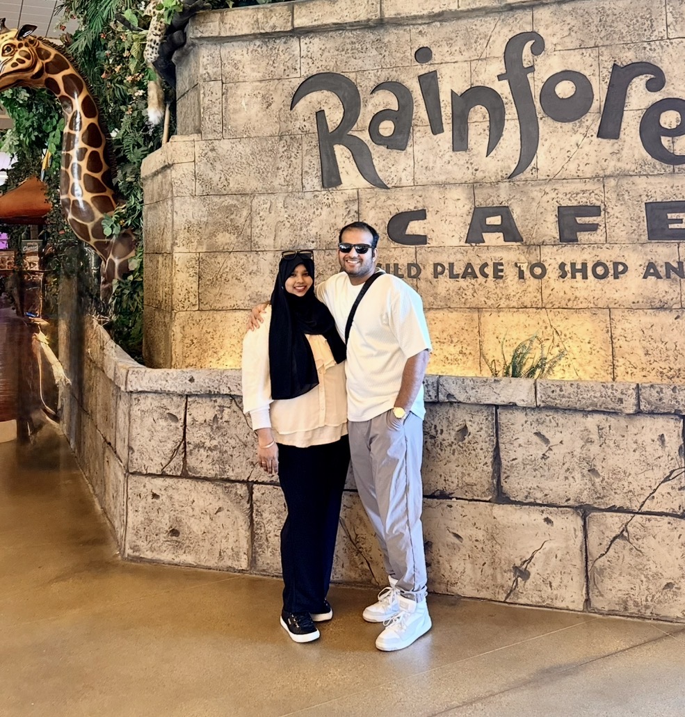 Two people stand in front of a stone wall with a sign that reads 'Rainforest'. A giraffe sculpture is visible on the left.
