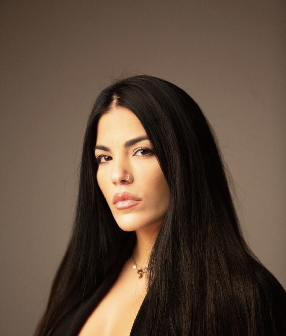 Woman with long black hair, neutral expression, wearing a necklace, against a plain background.