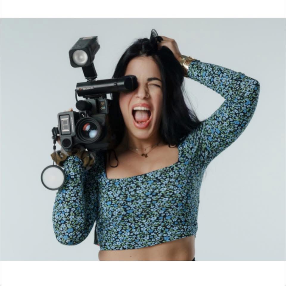Young woman with dark hair holding a camera, smiling with her mouth open, wearing a floral long-sleeve crop top.