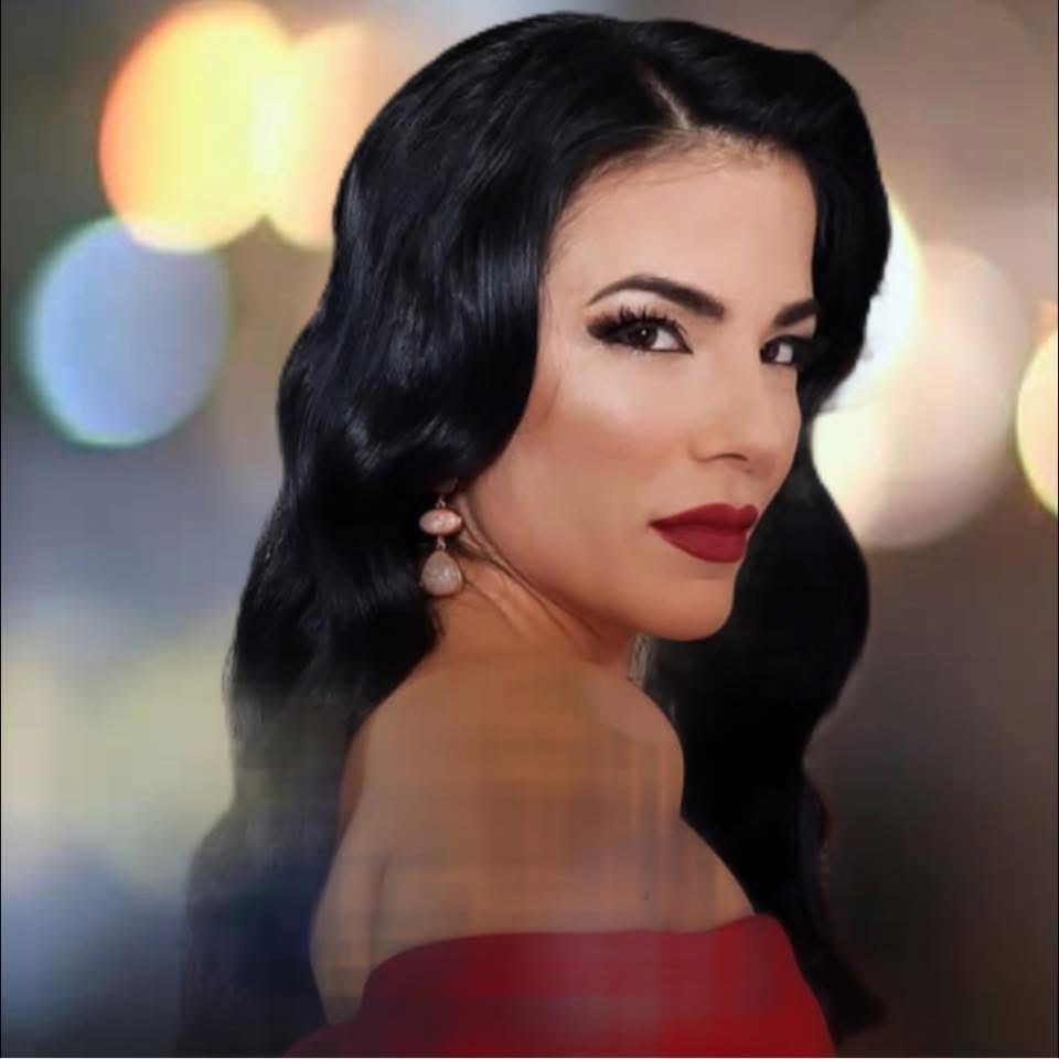 Woman with dark wavy hair, red lipstick, and earrings, looking over shoulder against blurred colorful background.