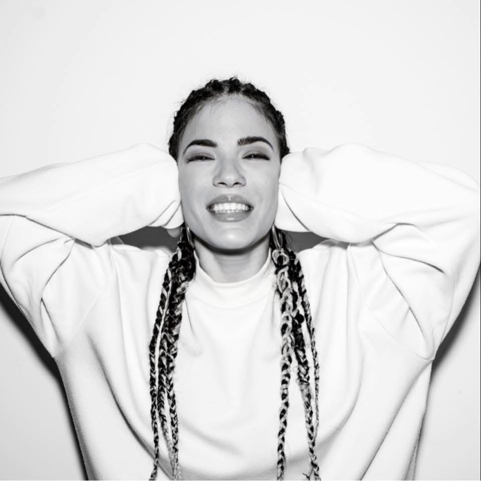 Woman with braided hair smiling and holding her head, wearing a loose sweatshirt, in front of a plain background.