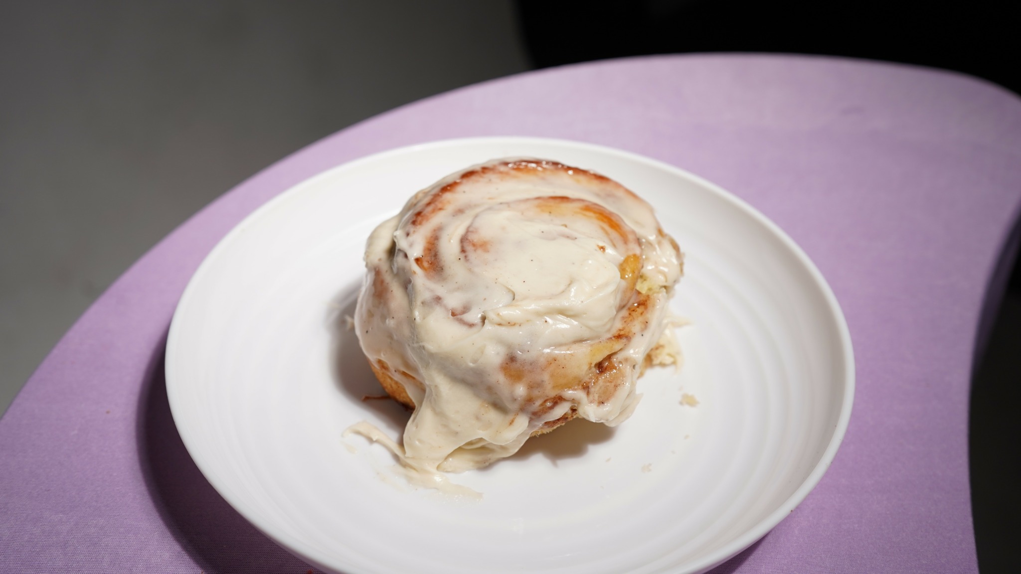 A cinnamon roll with icing on a white plate, placed on a purple surface.