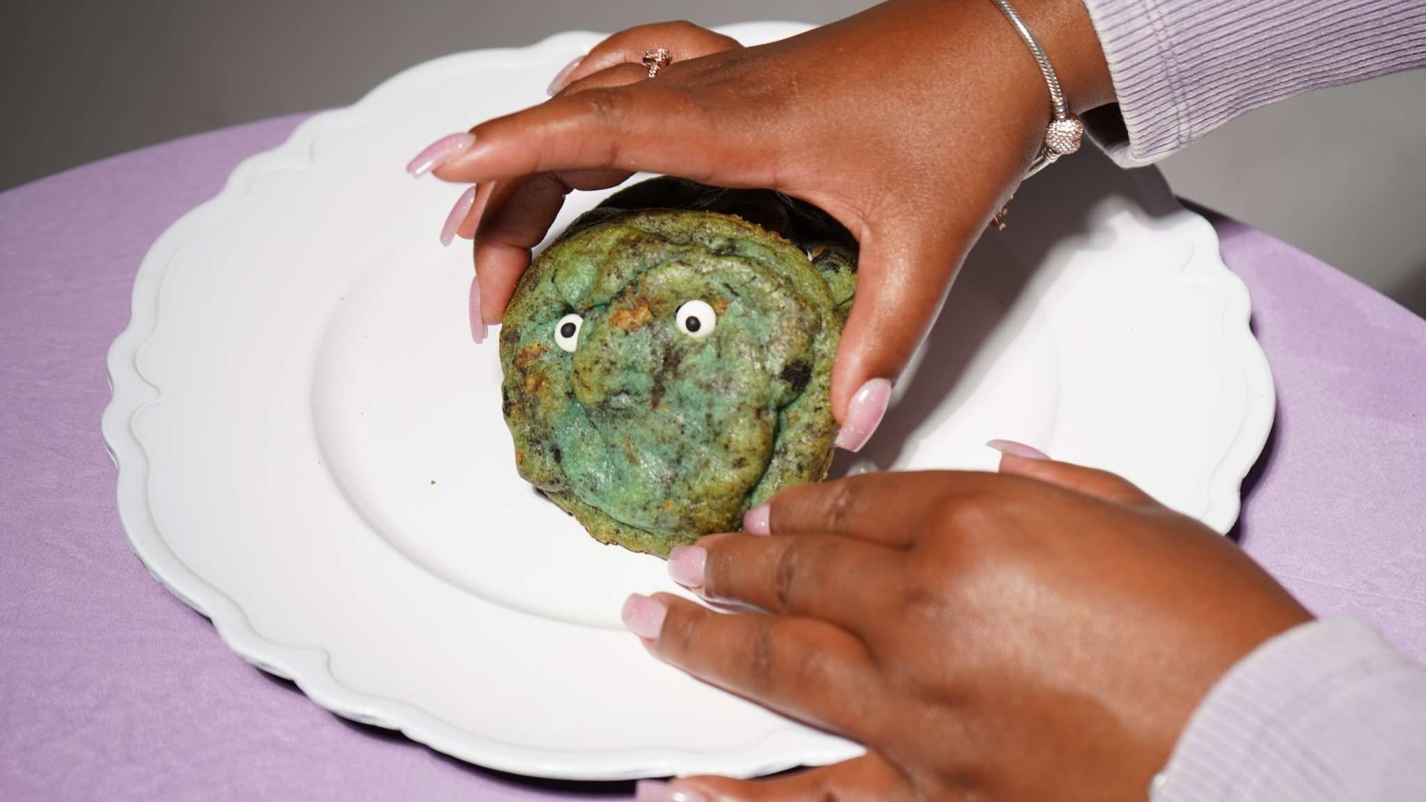 Person holding a green, textured object resembling a face on a white plate with a purple tablecloth.