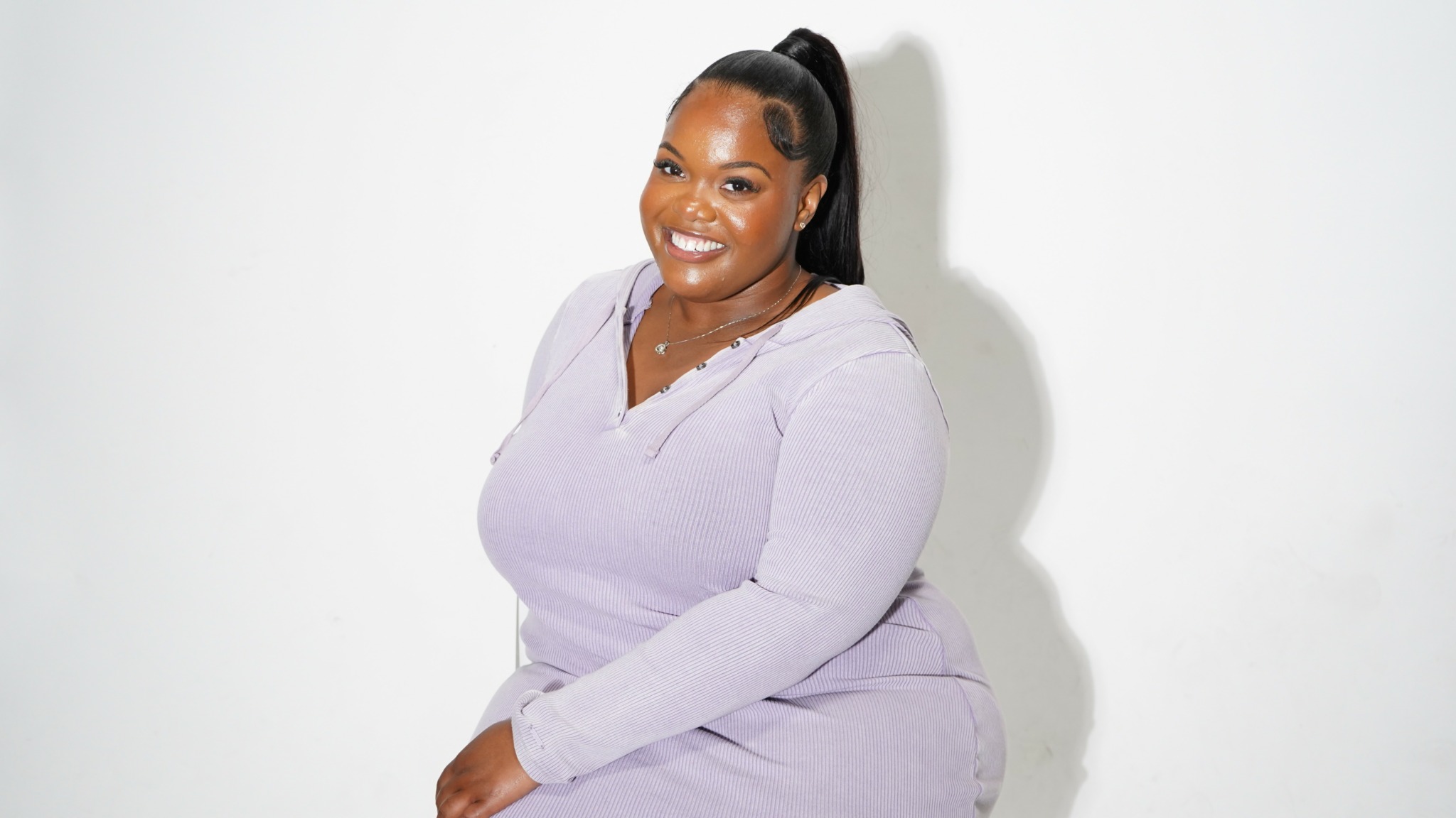 Smiling woman with long dark hair in a high ponytail, wearing a light purple outfit, sitting against a white background.