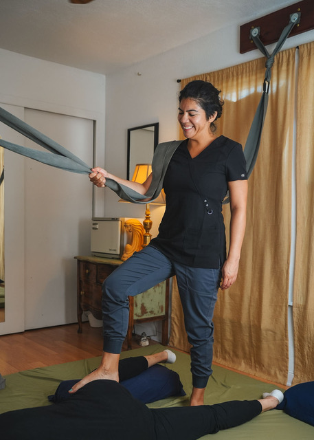 Woman smiling, standing on a bed with one foot on a pillow, holding a gray strap, in a room with yellow curtains.