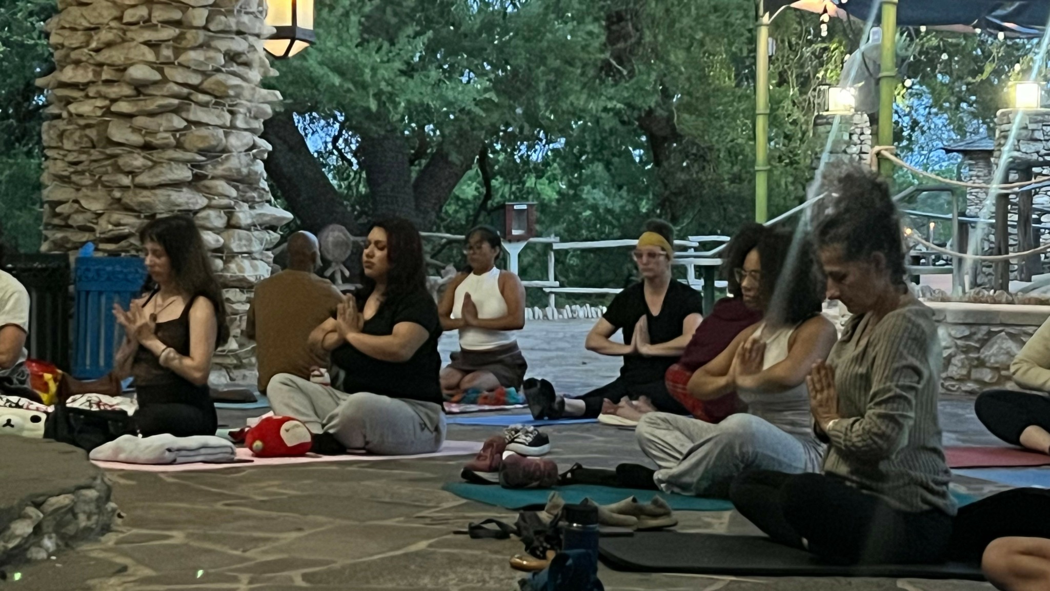 Group of people practicing yoga outdoors on mats, sitting cross-legged with hands in prayer position, surrounded by trees.