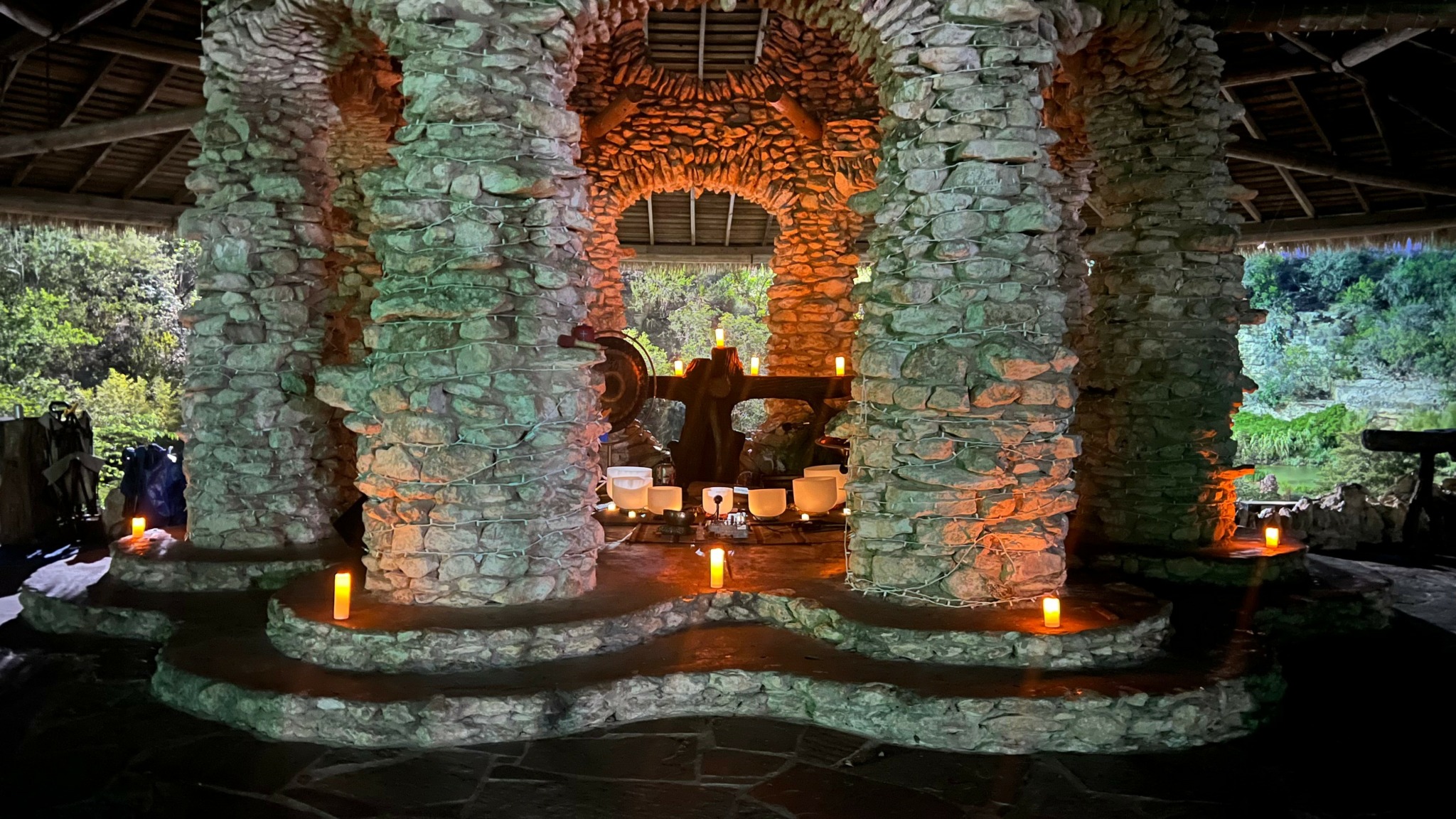 Stone fountain with three arches, candles around base, outdoor setting with trees in background.