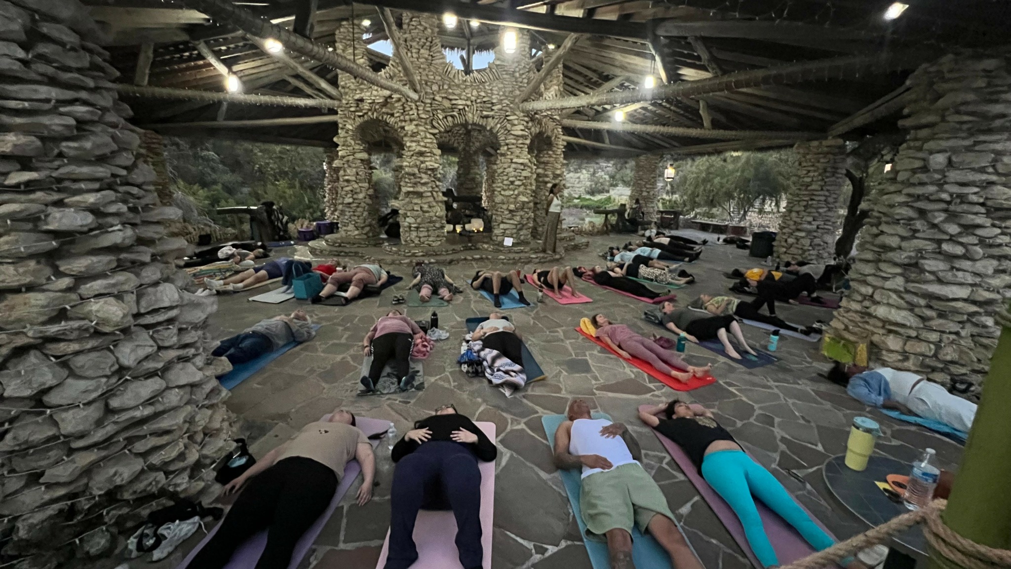 People participating in an outdoor yoga class under a stone structure with trees and stone pillars.