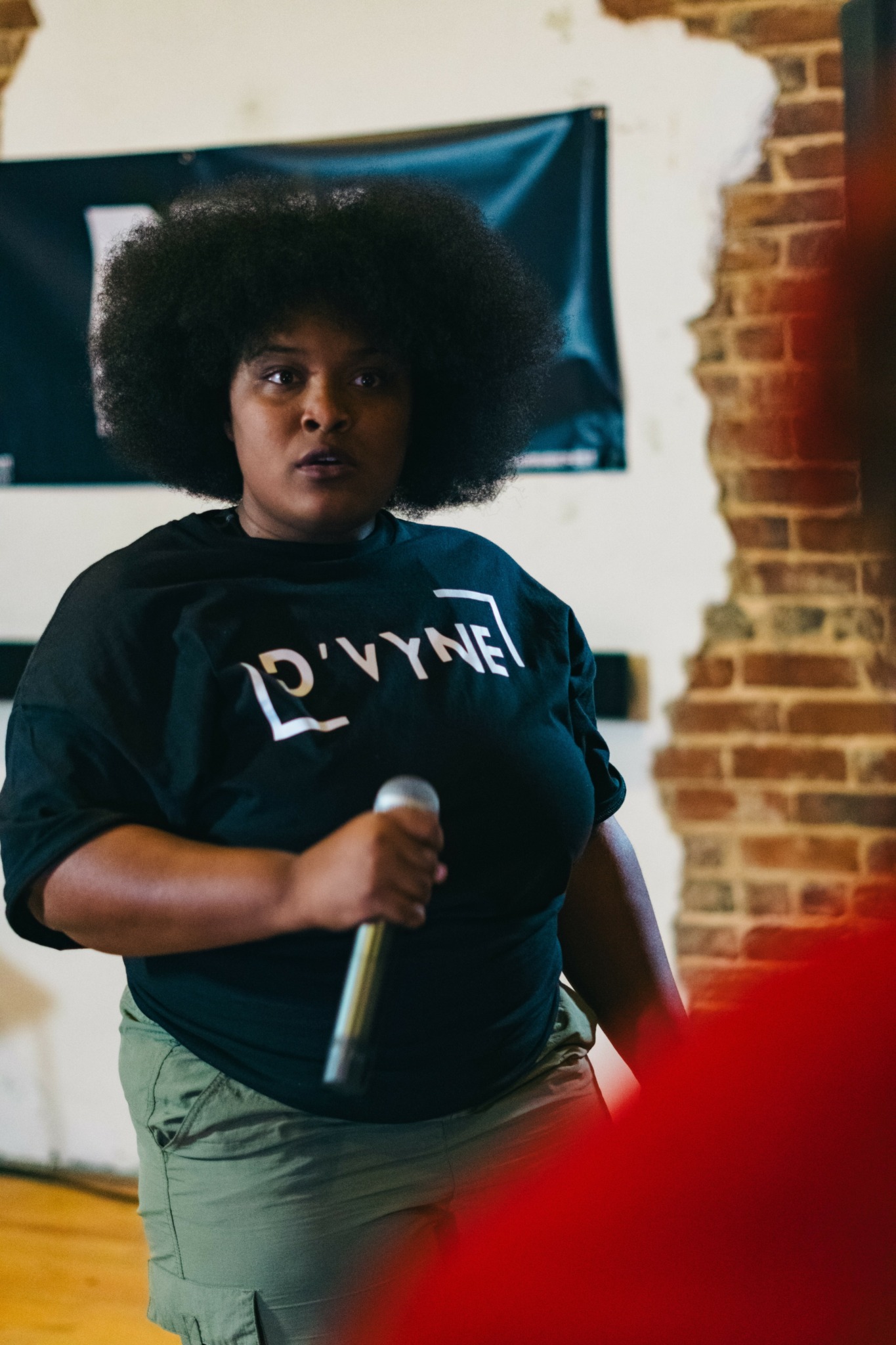 Woman with large afro holding a microphone, standing indoors with brick wall and banner behind her.