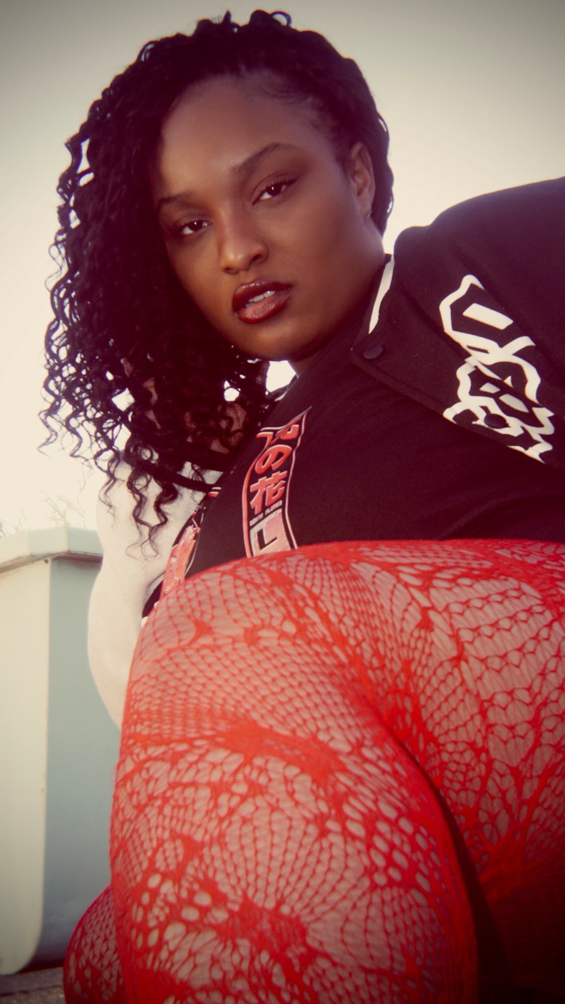 Woman with curly hair wearing black top and red lace stockings, looking at camera.