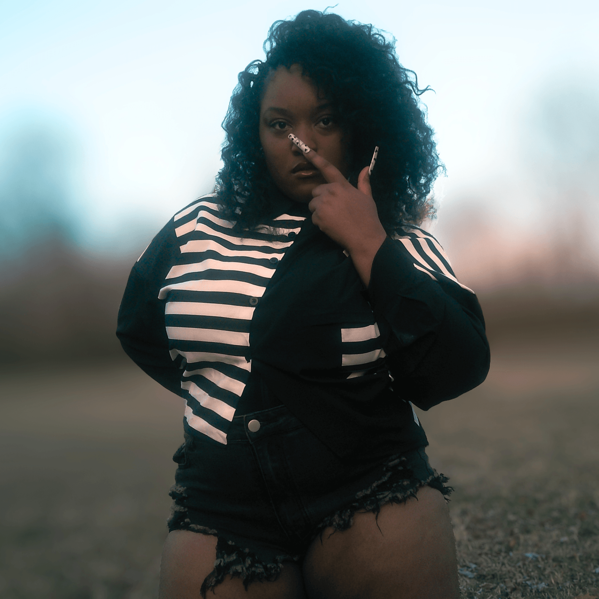 Woman with curly hair wearing a striped shirt and black jacket, standing outdoors with blurred background.