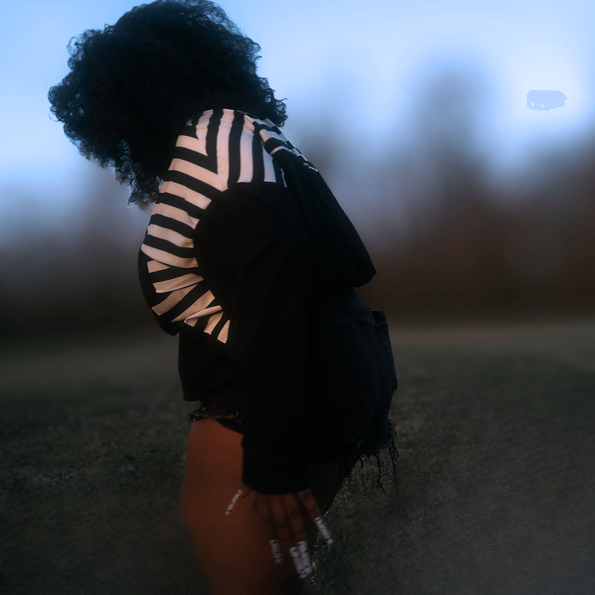 Person with curly hair wearing a striped shirt and black shorts, standing outdoors during dusk.