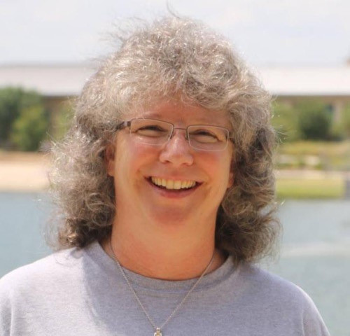 Smiling person with curly gray hair and glasses outdoors near water and a bridge.