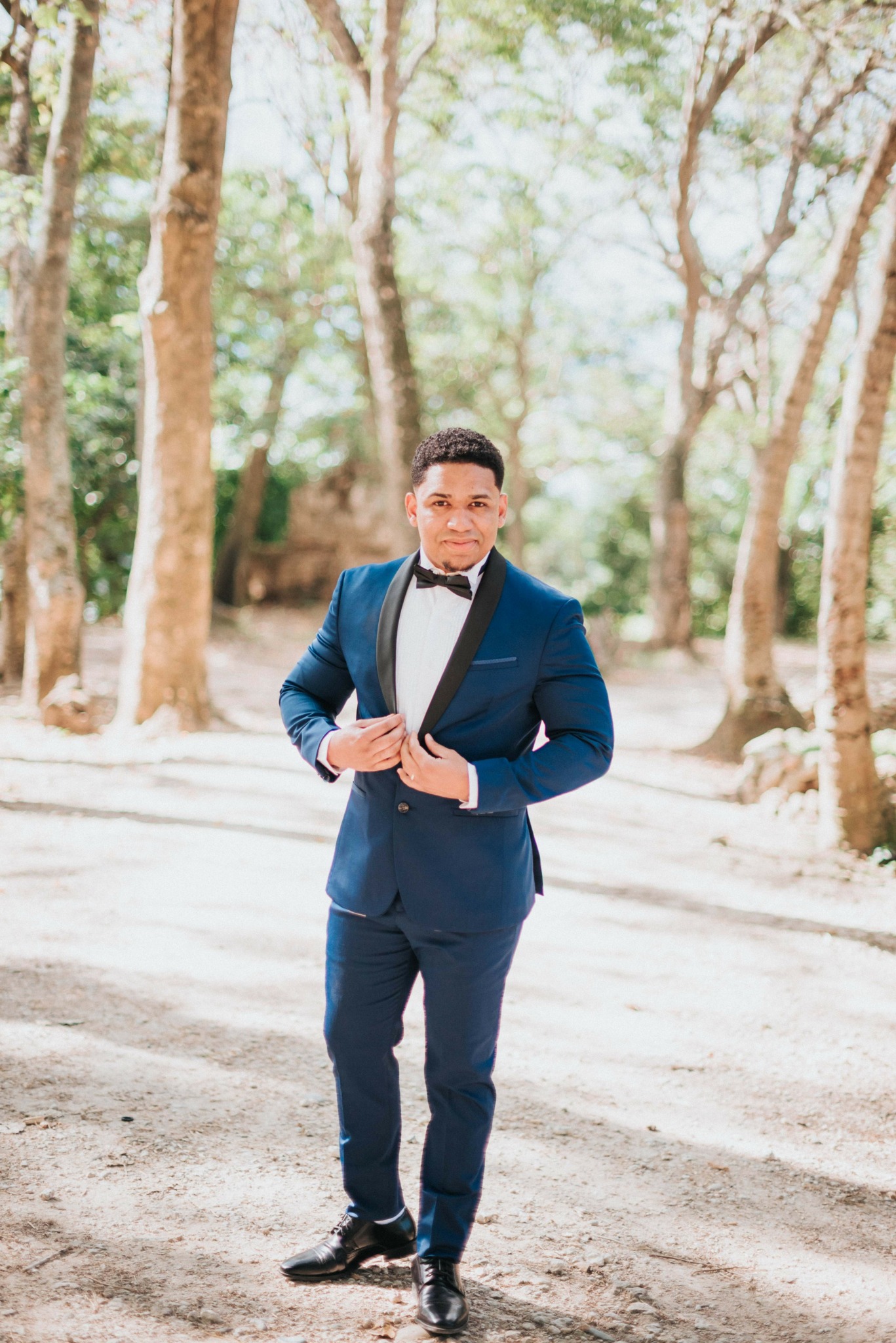 Young man in formal suit and bow tie walking outdoors among trees, sunlight filtering through leaves.