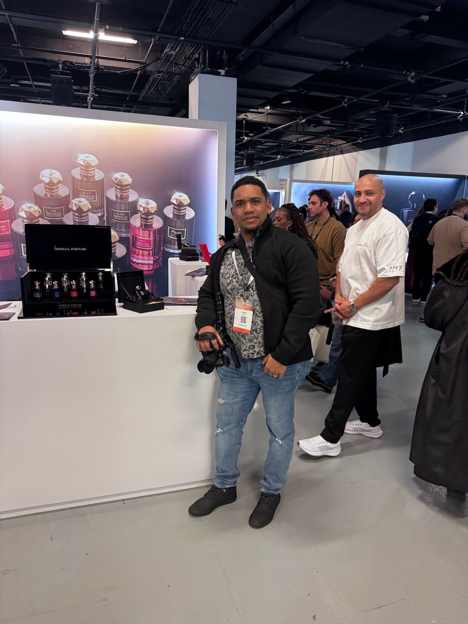 Two men standing in front of a display with bottles and a screen, one holding a camera, in an indoor setting.