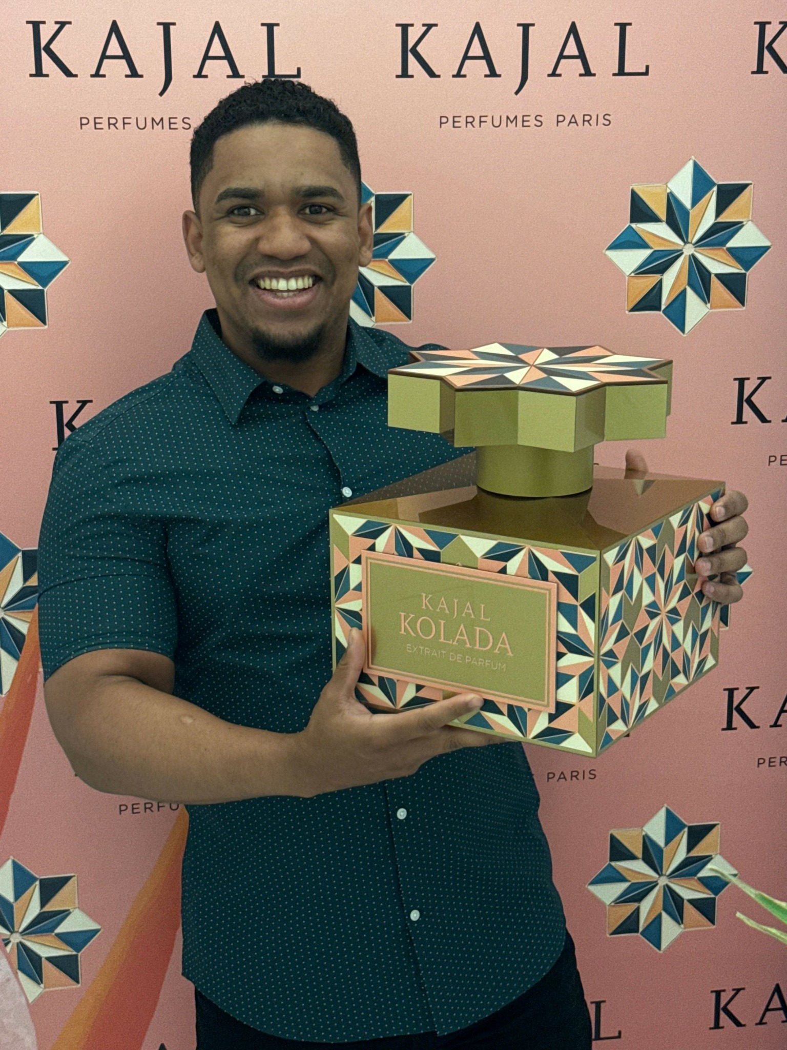 Person holding a box of Kaïal perfume in front of a pink background with geometric patterns.