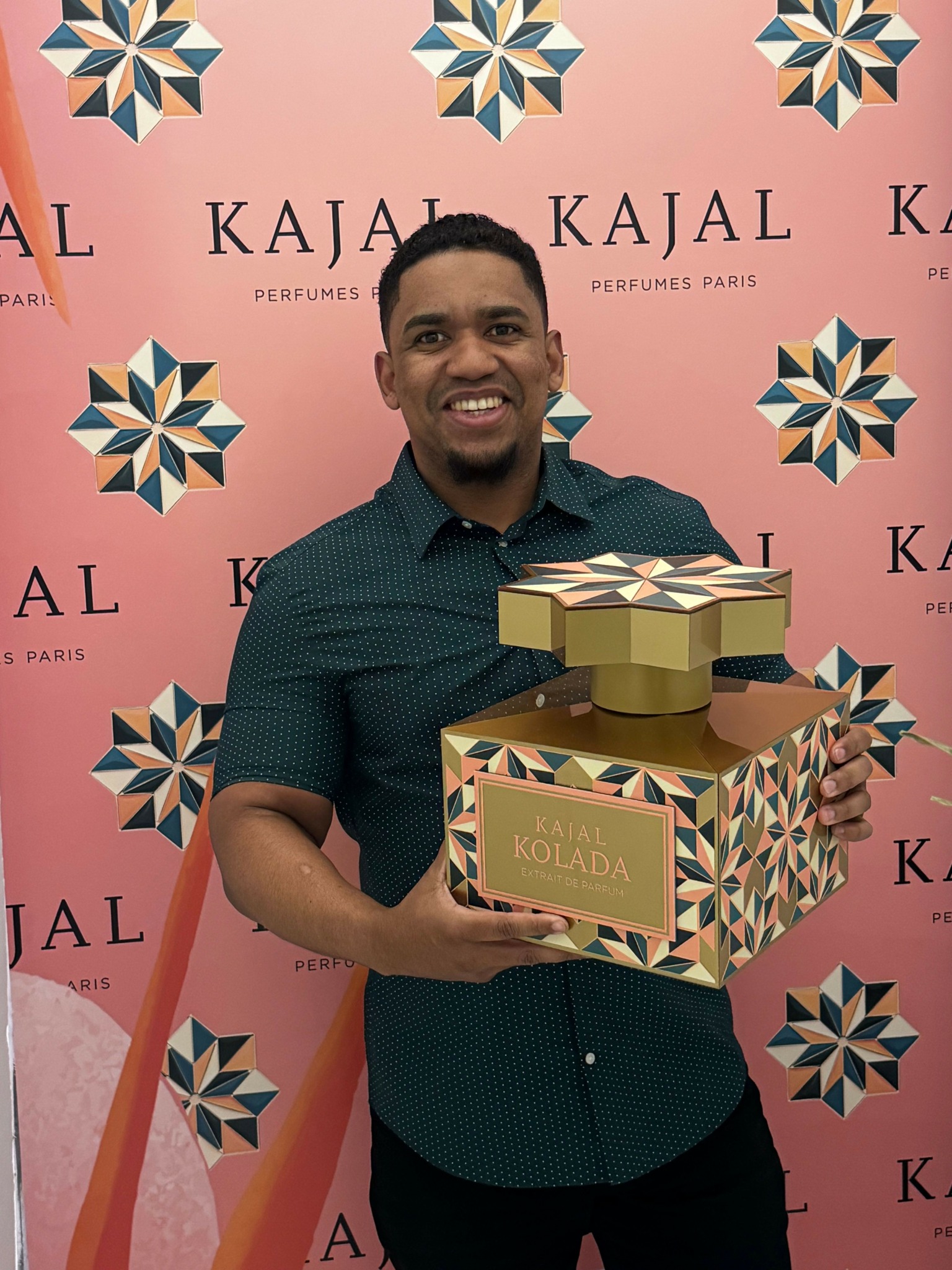 Person holding a trophy and a box, smiling, standing in front of a pink backdrop with logos and star patterns.