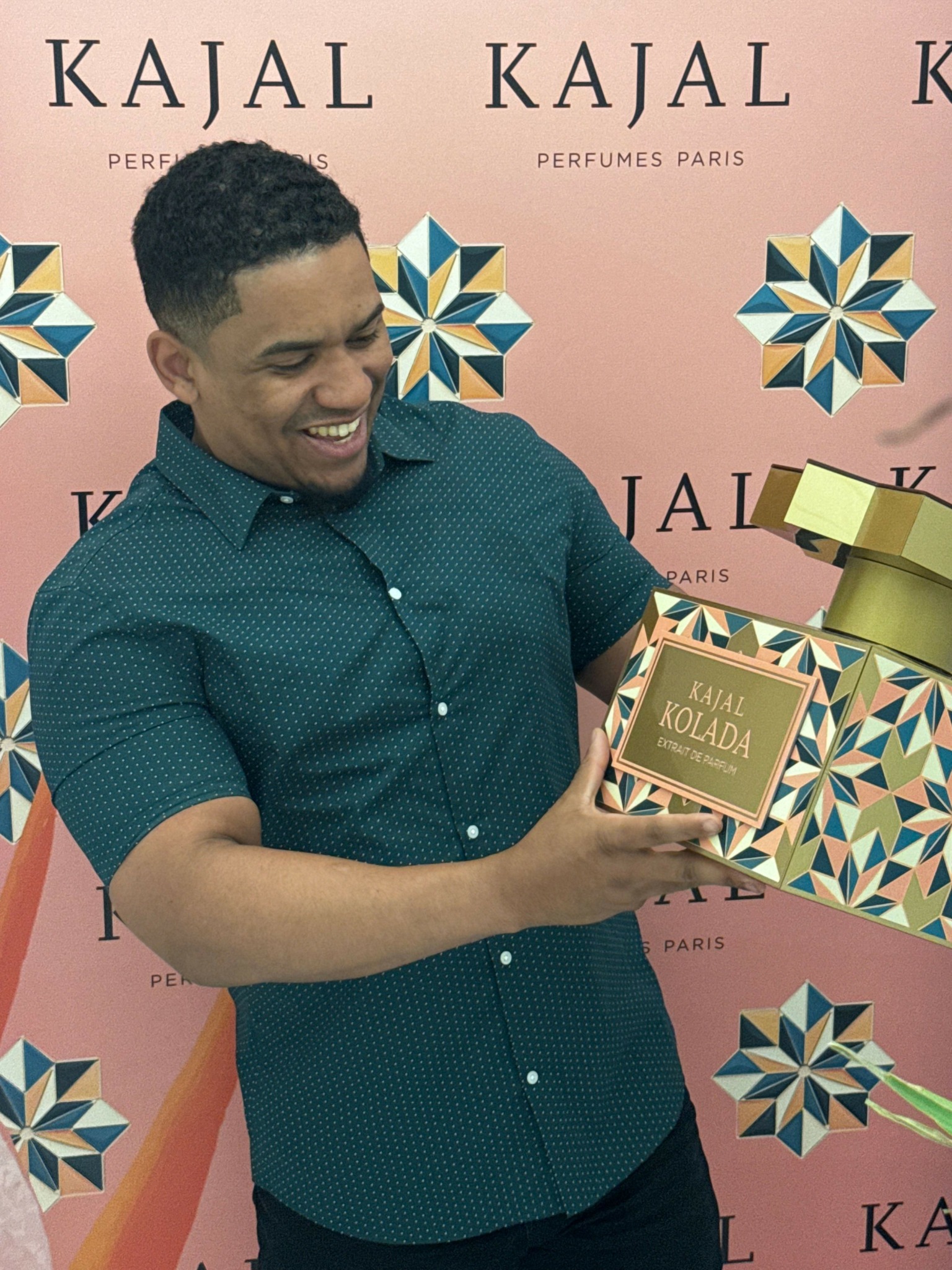 Person holding a gold box in front of a pink background with geometric patterns and the word 'KATAL' repeated.