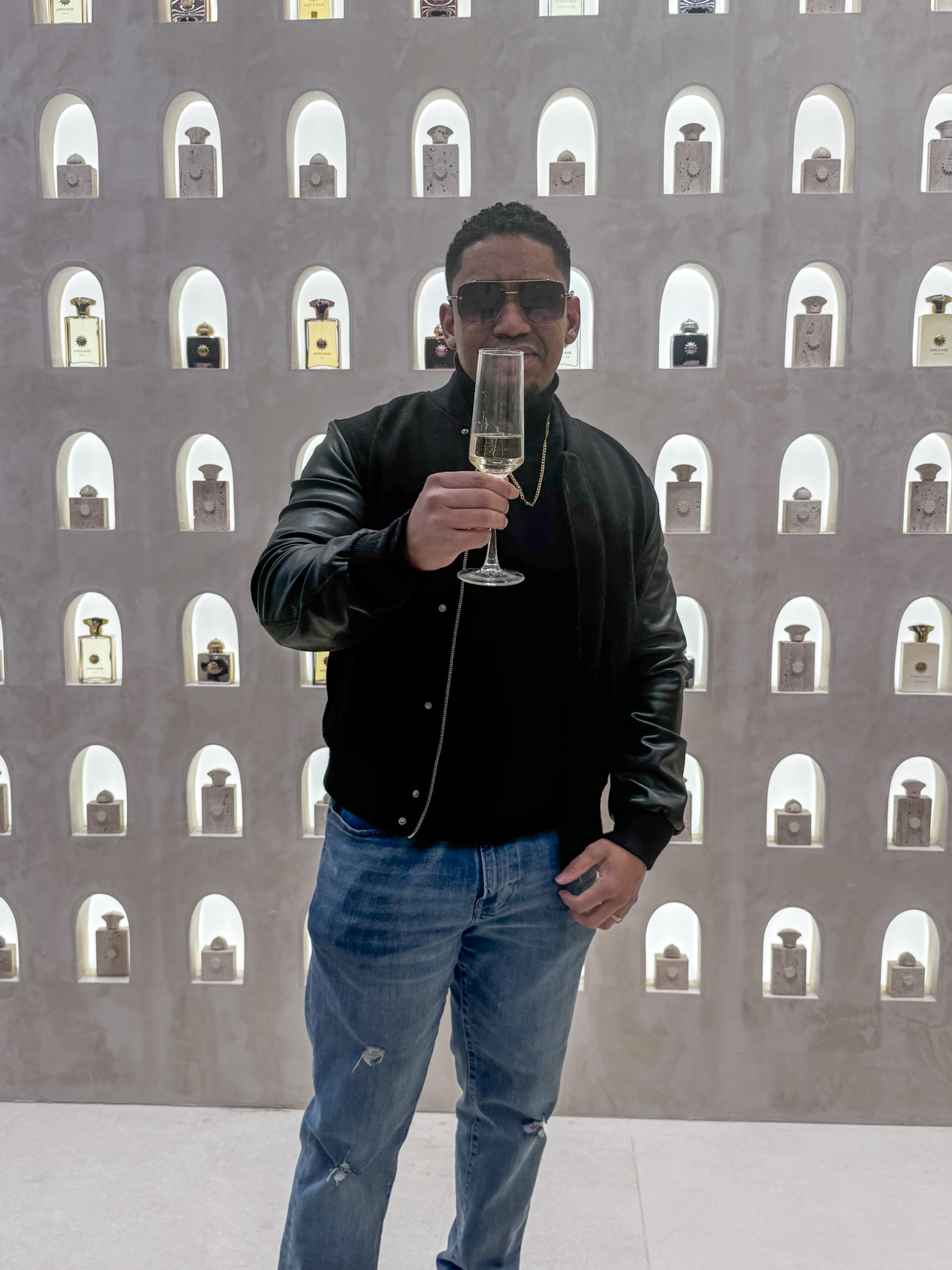 Man in black jacket and jeans holding a champagne glass in front of a wall with small arched niches.