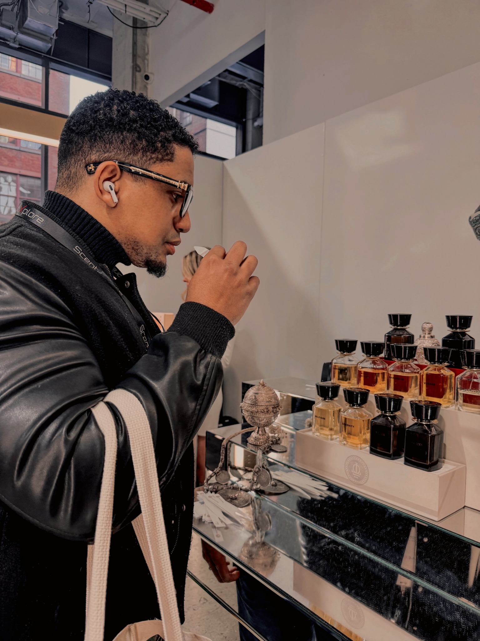 Person with short curly hair, glasses, black jacket, looking at perfume bottles on display.
