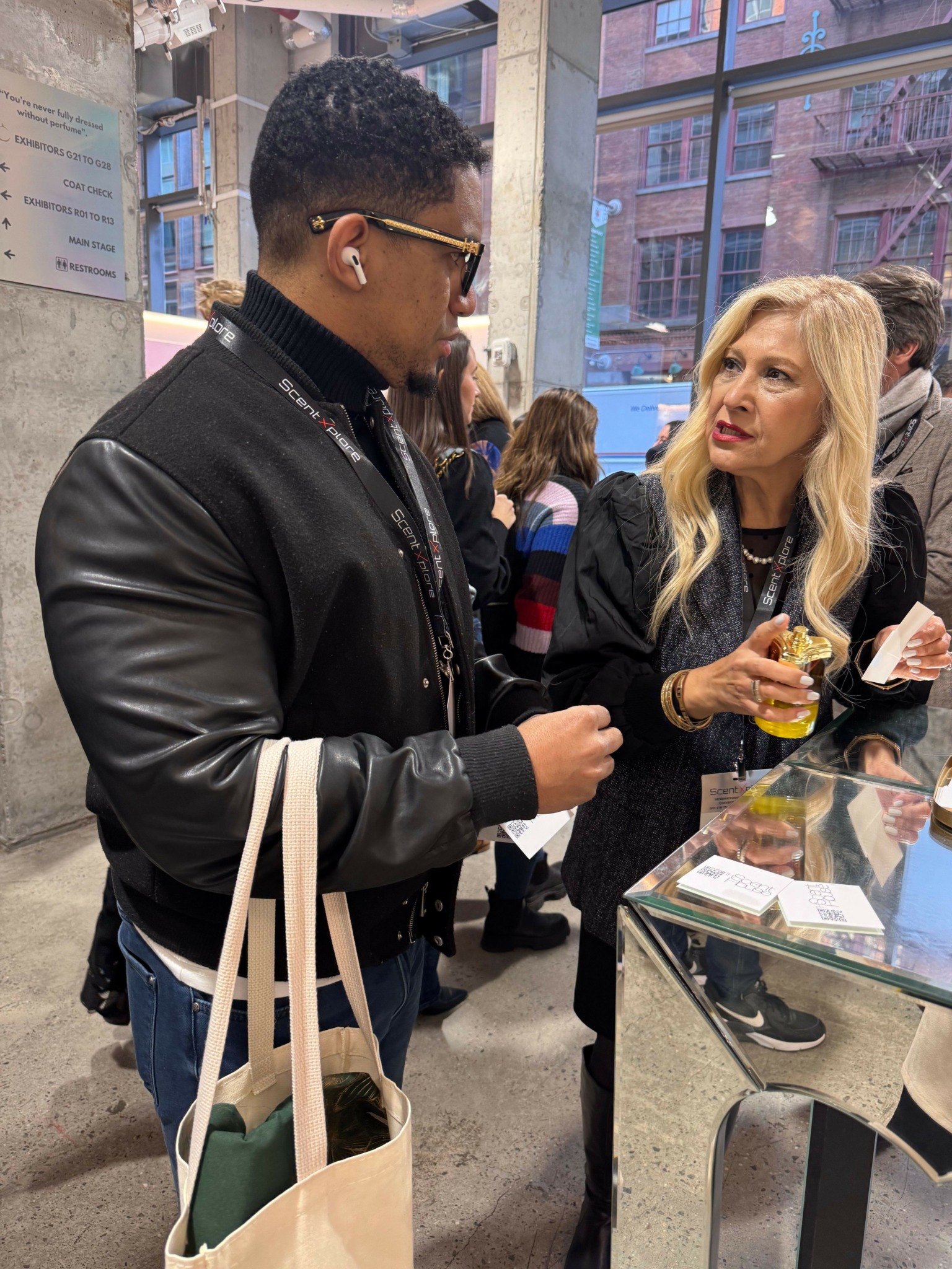Two women standing at a table, one holding a yellow drink, in a busy indoor setting with large windows.