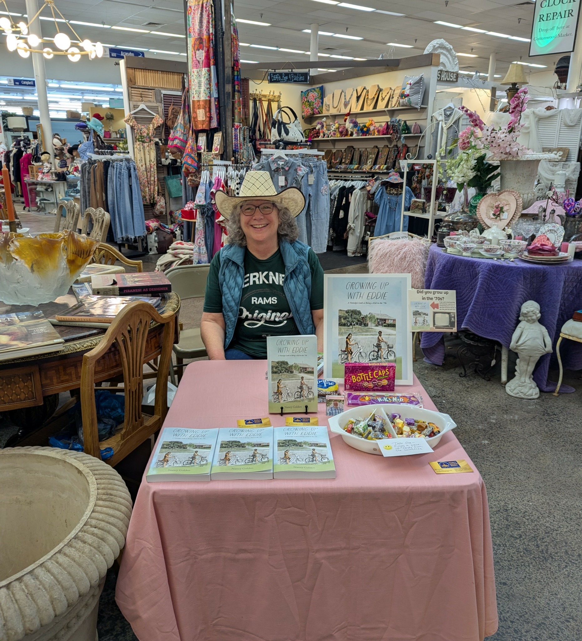 Woman with a large hat and glasses sitting at a table with books and a bowl, in a store with various items.