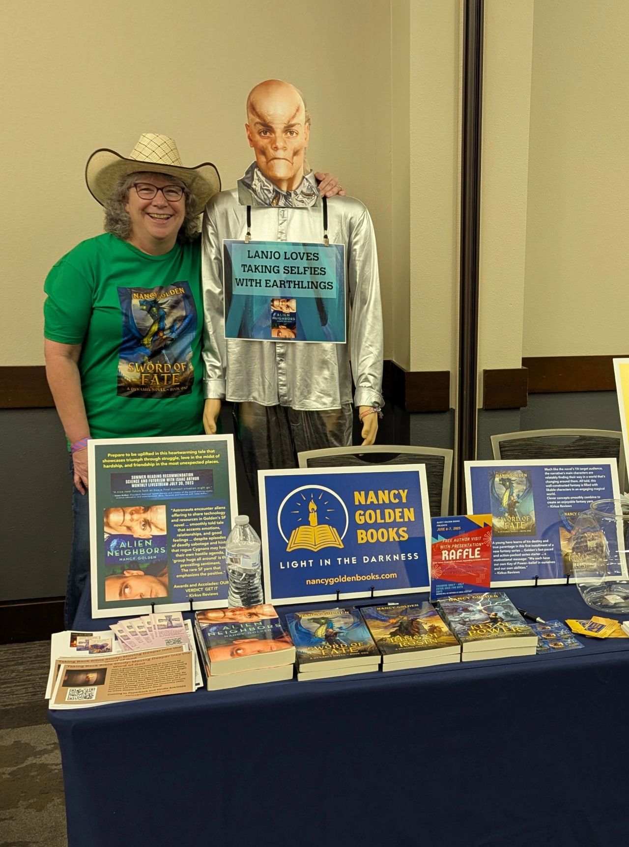 Woman in green shirt and a large hat standing next to a puppet with a bald head and suit, behind a table with books and displays.
