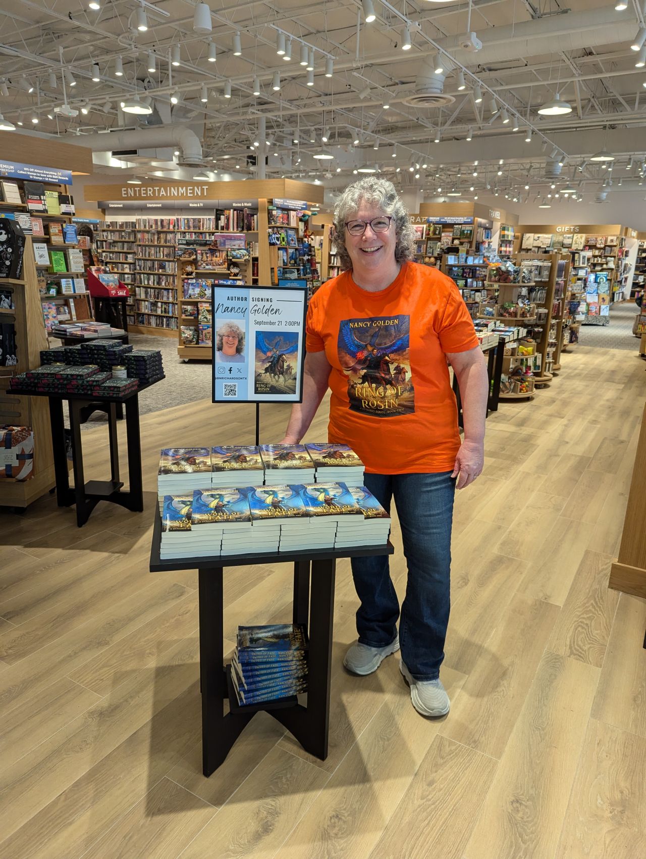 Woman with glasses and curly hair wearing an orange T-shirt and jeans standing next to a display table in a bookstore.