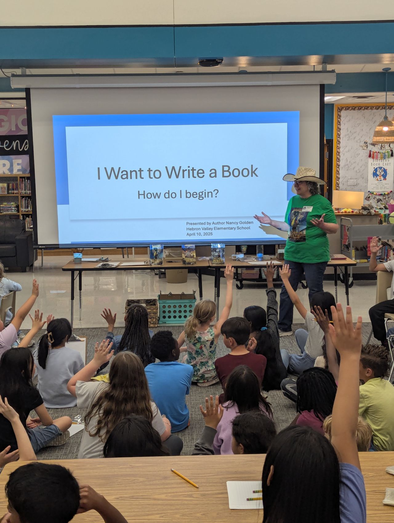 Teacher standing in front of a classroom with students raising hands, presentation slide on screen about writing a book.