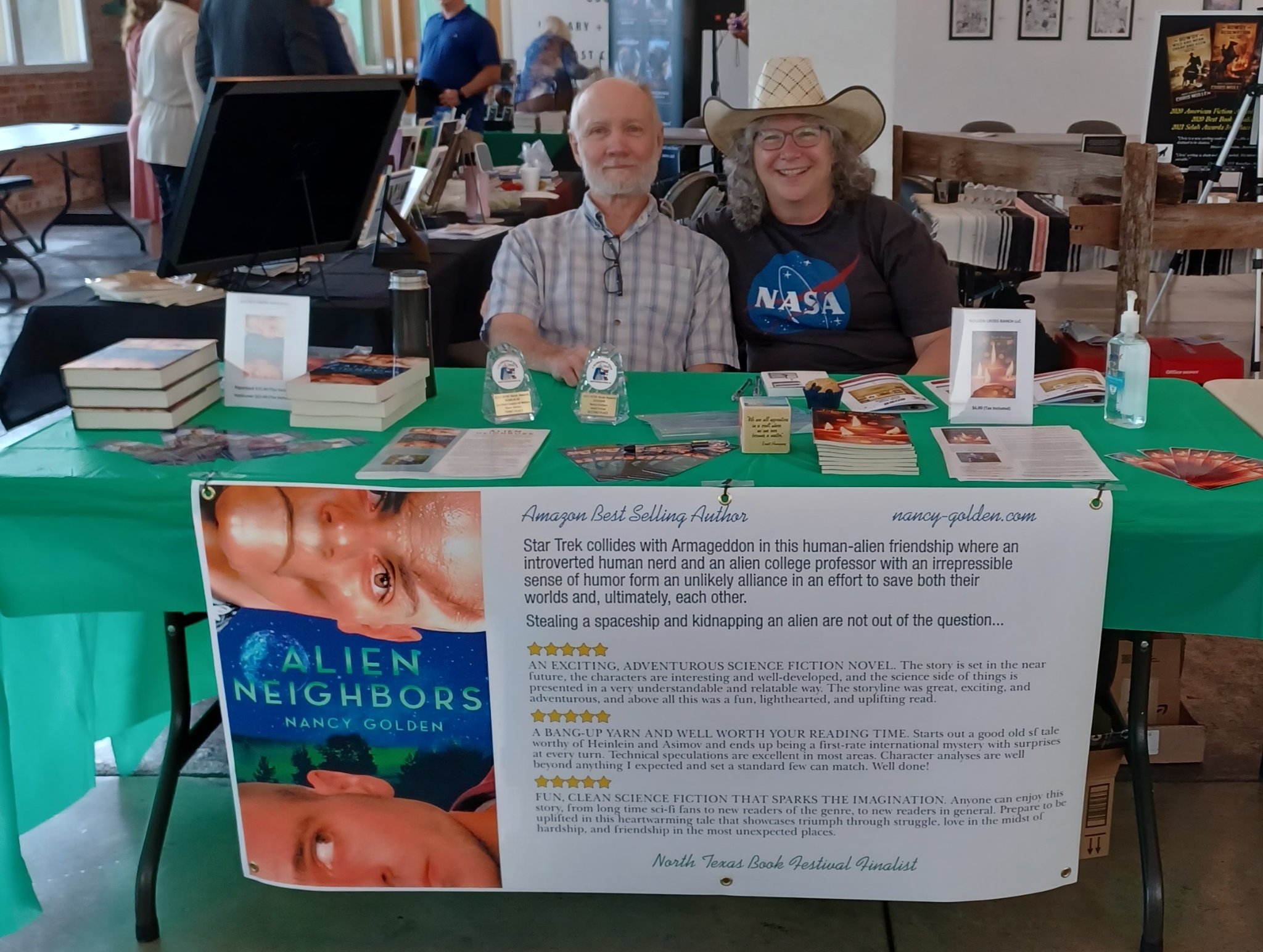 Two people sitting behind a table with books and a large informational sign, in an indoor setting.